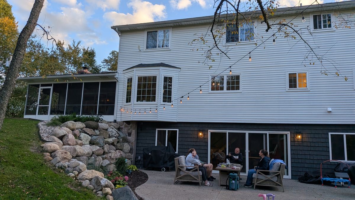 Back of the house at dusk: white siding, a bay window, string lights, and the walkout patio with family sitting outside
